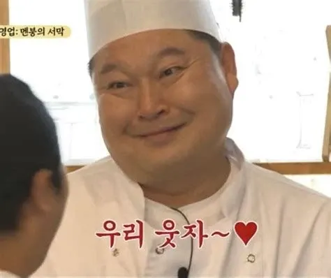 요리사 복장에 흰 모자를 쓴 남자가 맞은편 사람을 바라보며 어색하게 웃고 있고, 화면 아래에 분홍색 글씨로 “우리 웃자~♥”라는 자막이 깔린 예능 프로그램 장면